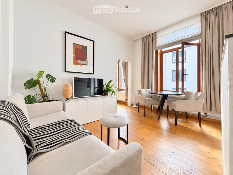 Modern living room with light wooden flooring, large windows, and a balcony. Features a sofa with a zebra-patterned throw, a round coffee table, two armchairs, a TV on a white cabinet, and decorative plants.