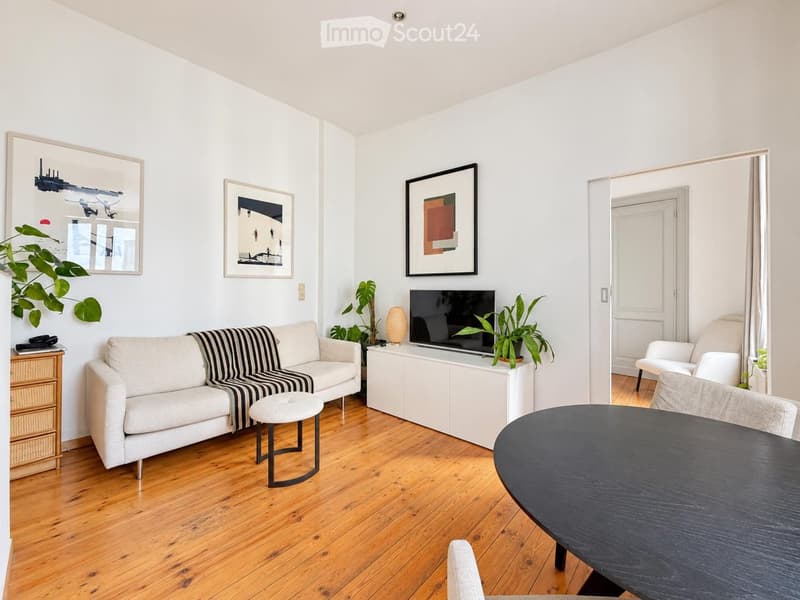 A modern living room with a white sofa, a small round table, a TV on a white cabinet, and several potted plants. The room features light wooden flooring and white walls adorned with several picture frames.