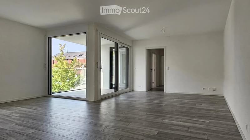Empty room with wooden flooring, large sliding glass doors leading to an outdoor area, and a doorway to another room.