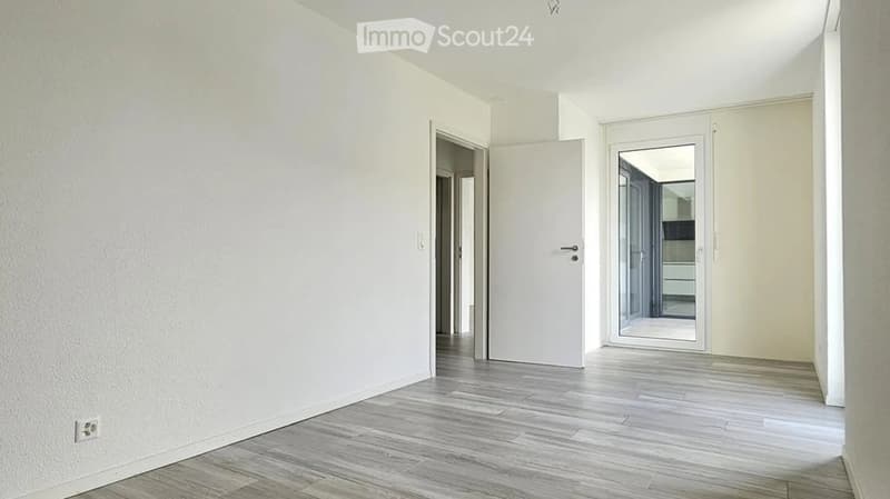 Empty hallway with light-colored walls, wooden flooring, and a large mirror on one wall. There are two closed doors and an open doorway leading to another room.