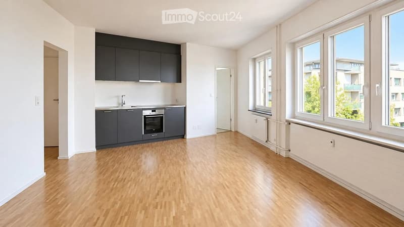 Empty room with light wooden flooring, white walls, and large windows. Kitchen area with cabinets and appliances is visible on the left side.