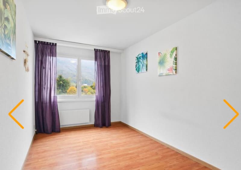 Empty room with wooden flooring, white walls, large window with purple curtains, and two decorative paintings on the wall.