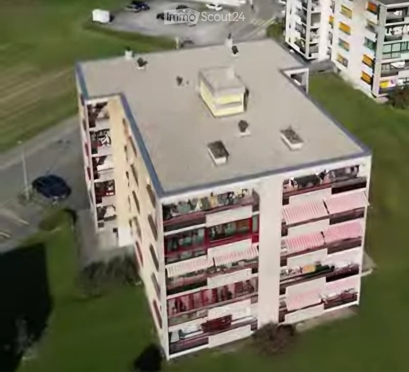 A multi-story residential building with balconies and a flat roof, surrounded by greenery and other structures.