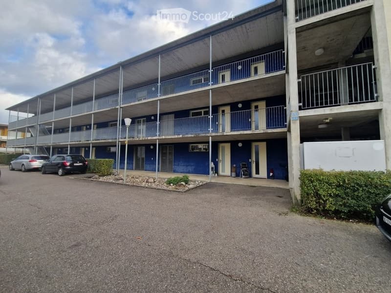 Multi-story apartment building with blue exterior, balconies, and parking area.