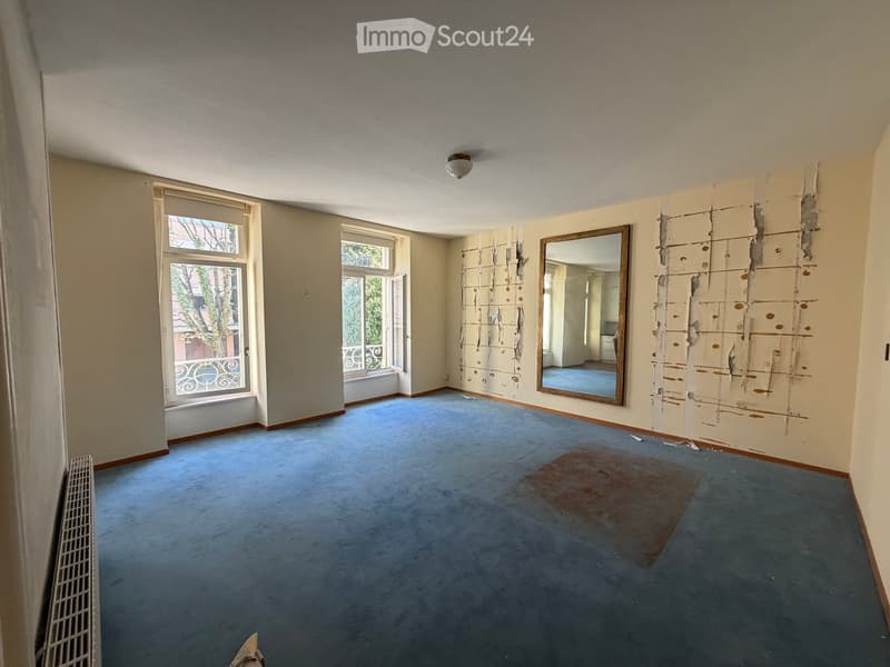 Empty room with blue carpet, exposed wall patches, large windows, and a mirror on one wall.