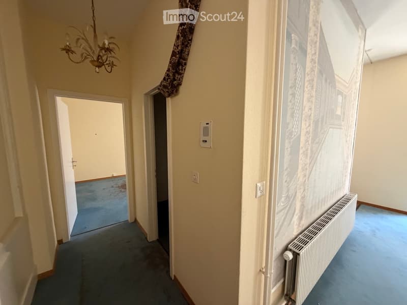 A narrow hallway with light-colored walls, blue carpeting, and a radiator on the right side. A chandelier hangs from the ceiling, and there are two open doors leading to other rooms.
