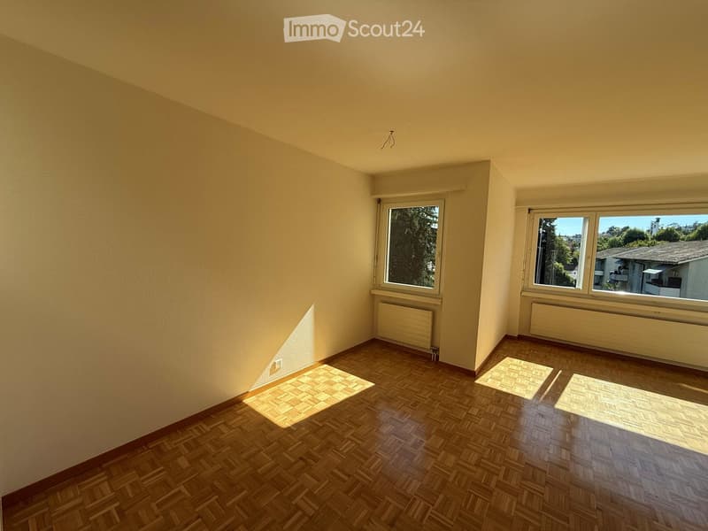 Empty room with wooden flooring, light-colored walls, and large windows allowing natural light to enter. Radiators are visible beneath the windows.