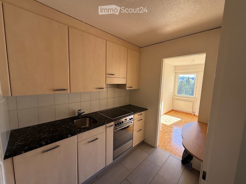 Modern kitchen with light-colored cabinets, granite countertop, stainless steel sink, stove, and oven. Adjacent room features wooden flooring and a small table.