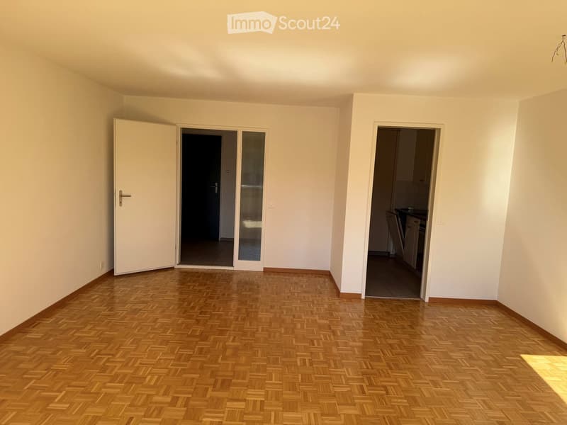 Empty room with wooden parquet flooring, white walls, and open doors leading to a balcony and a kitchen area.