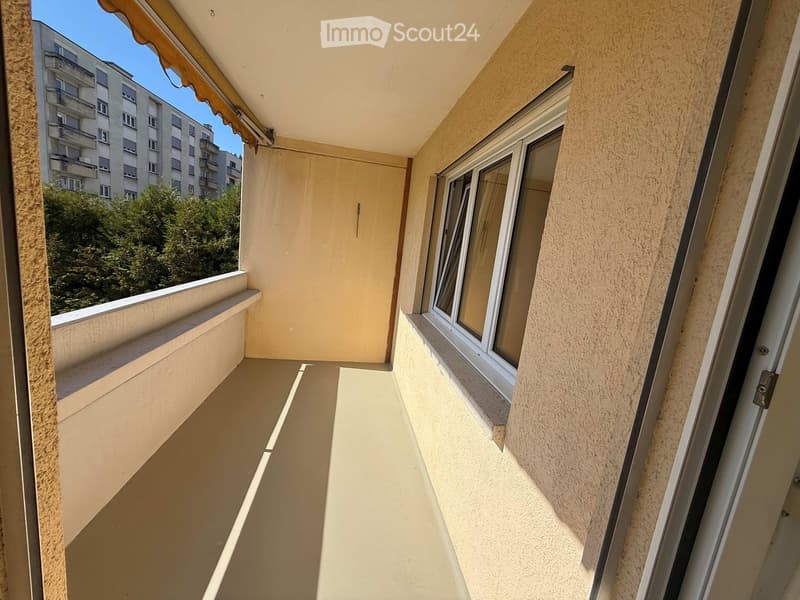Balcony with beige walls, a window with white frame, and an awning above. The view shows a building and trees in the background.
