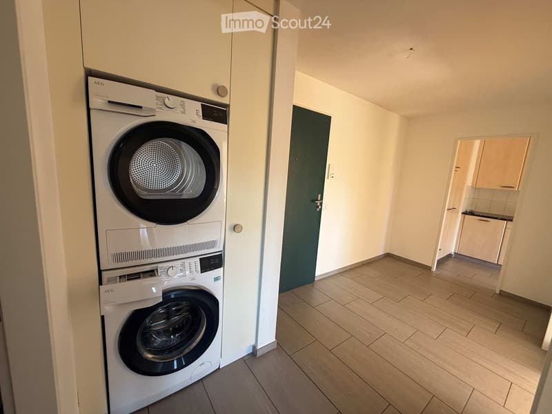 Hallway with tiled floor, white walls, a green door, and a stacked washing machine and dryer unit on the left side.
