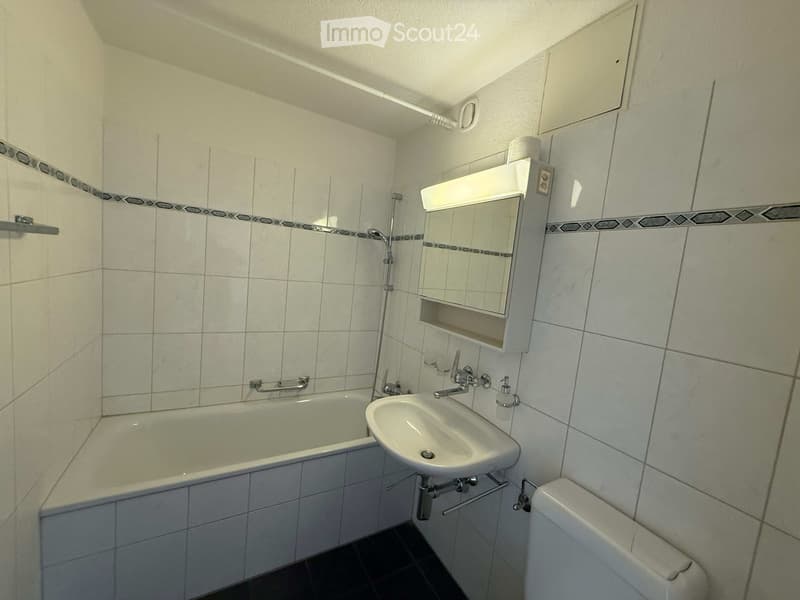 A small bathroom with a bathtub, sink, toilet, and a mirror above the sink. The walls are tiled with a decorative border, and there is a showerhead and faucet above the bathtub.