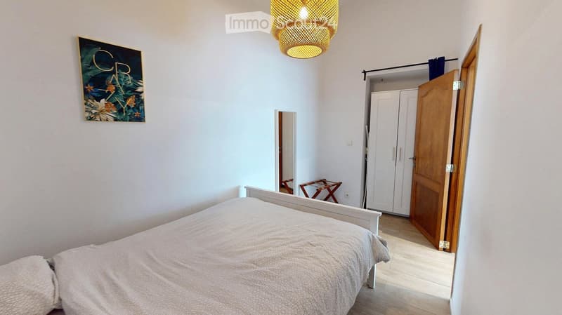 A modern bedroom with a white bed, a wooden wardrobe, a mirror, a small wooden stool, a decorative picture frame, and a stylish wicker pendant light.