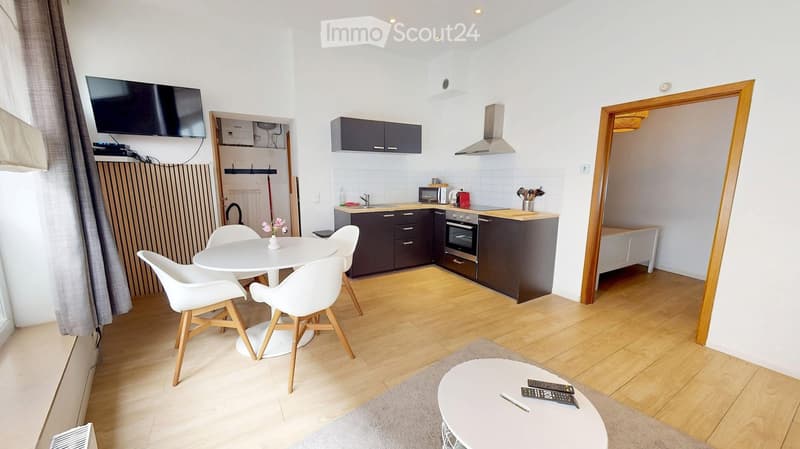 A modern, open-plan living and dining area with a kitchen. Features include a white dining table with four chairs, a small round coffee table with remote controls, a flat-screen TV mounted on the wall, and a compact kitchen with dark cabinets, a stainless