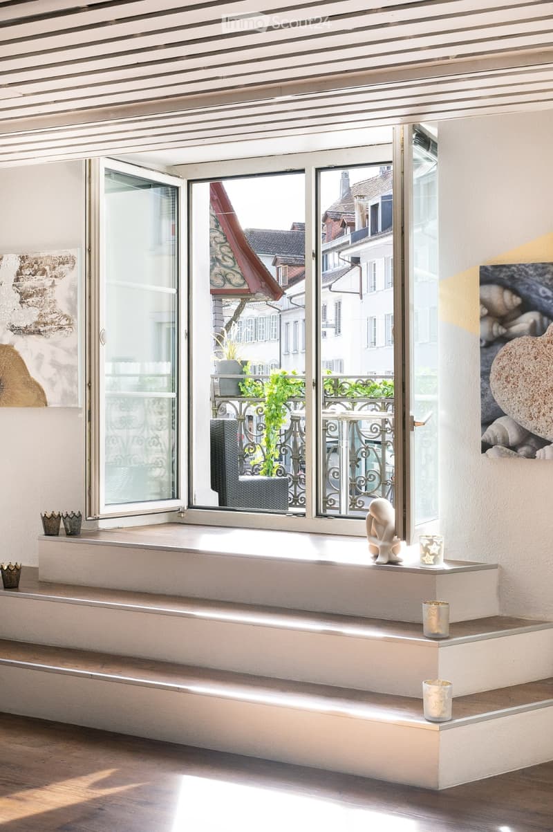 Bright room with large windows offering a view of the street, featuring a white table with decorative items including a statue and candle holders.