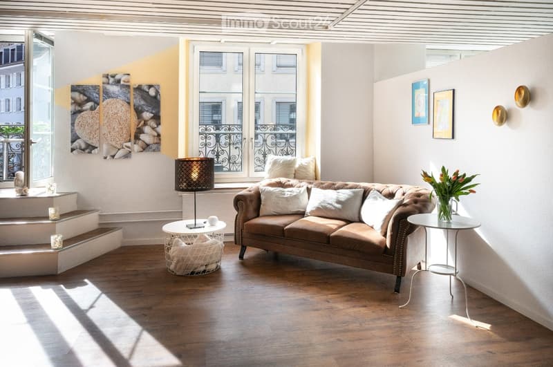 Modern living room with a brown leather sofa, white throw pillows, a round coffee table, a side table with a vase of flowers, a floor lamp, and decorative artwork on the walls.