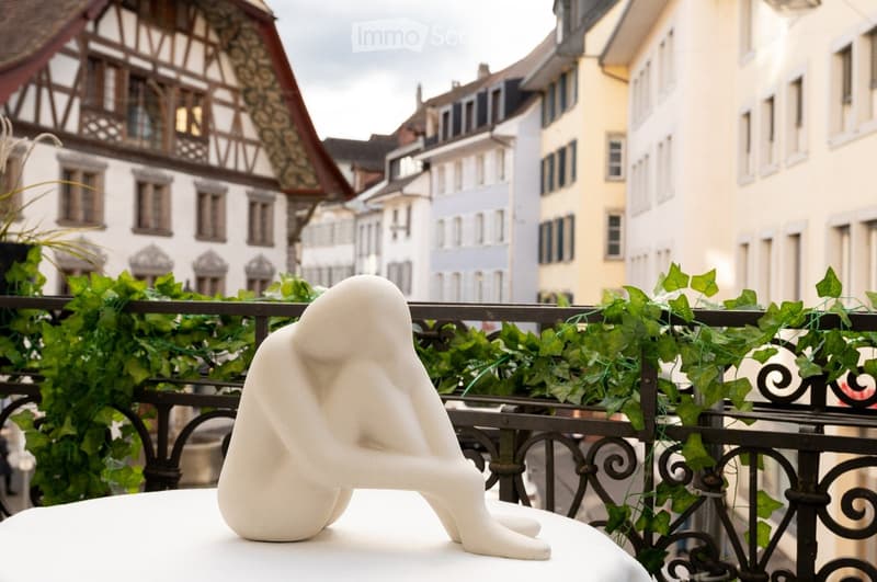 Balcony with a white table, a white sculpture, and green ivy climbing on the railing, overlooking a historic town with traditional buildings.