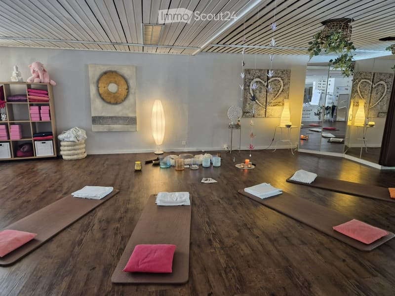 A spacious yoga studio with wooden floors, yoga mats, pillows, shelves with folded towels, a decorative wall art, and a mirror. The room is well-lit with hanging lights and has a calm, serene atmosphere.