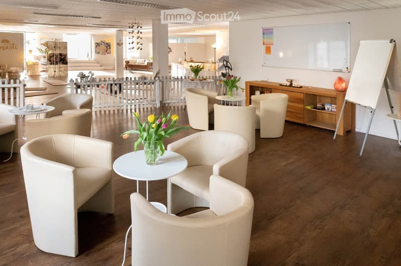 Modern, well-lit lounge area with several white chairs, small round tables, and vases with flowers. A large screen is visible on the right side.