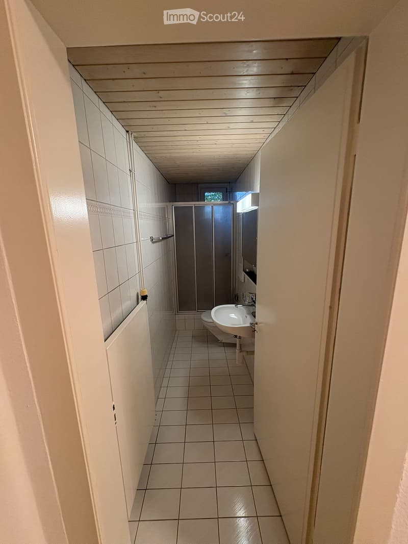 A narrow hallway with tiled walls and floor, leading to a small bathroom area with a sink, toilet, and shower stall.