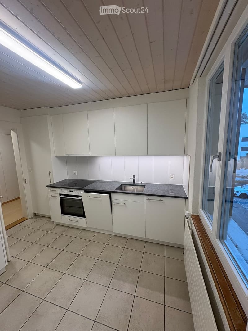 Modern kitchen with white cabinets, black countertop, built-in oven, sink, and faucet. The space features tiled flooring and large windows.