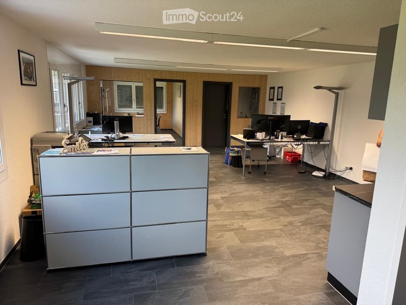 Office space with reception desk, two desks with computers, office chairs, printer, and framed pictures on the walls.