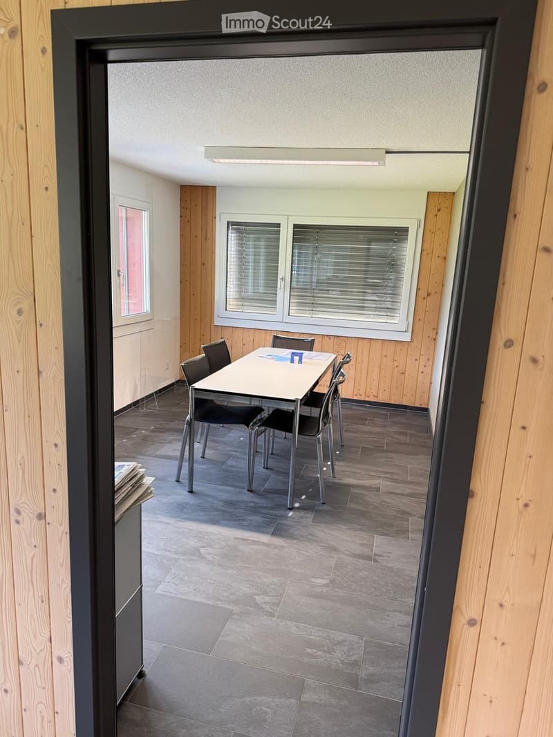 A small, furnished room with a table and chairs, wooden walls, and large windows with blinds.