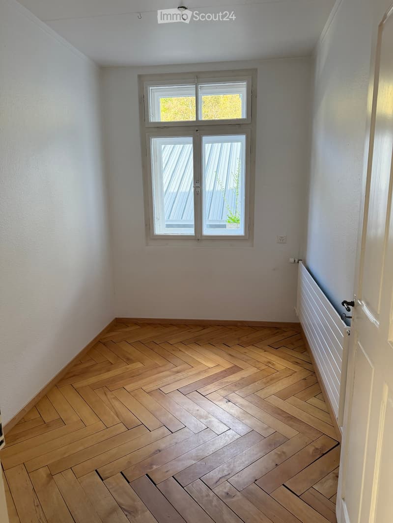 Empty room with wooden herringbone flooring, white walls, and a large window with double glass panels. A radiator is visible on the right side.