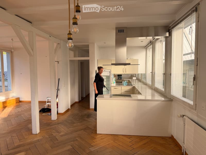 Bright, open kitchen with white cabinets, a central island, and wooden herringbone flooring. Large windows provide natural light, and modern appliances are visible.