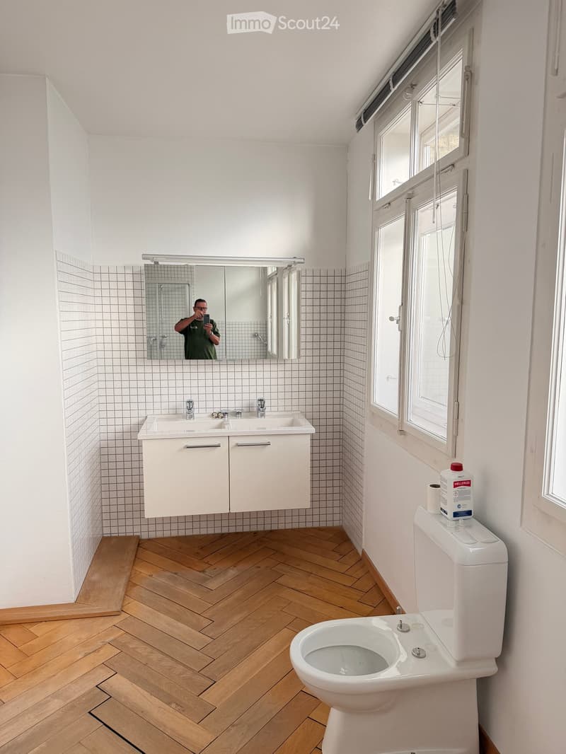 Modern bathroom with white tiled walls, wooden herringbone floor, double sink with mirrored cabinet, toilet, and window with blinds.