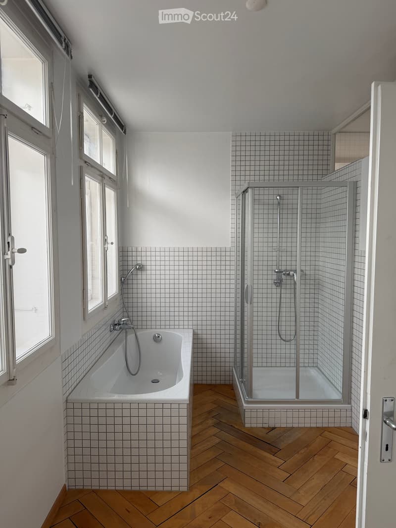 Modern bathroom with a white tiled wall, a wooden herringbone floor, a bathtub, and a glass-enclosed shower area.