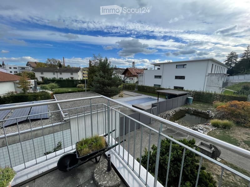 Balcony overlooking a landscaped garden area with a small pond, solar panels, and surrounding residential buildings.