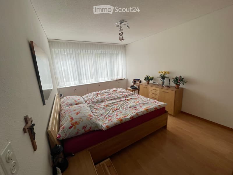 Cozy bedroom with wooden flooring, a bed with floral bedding, a nightstand, a chair, and a cabinet with decorative items.