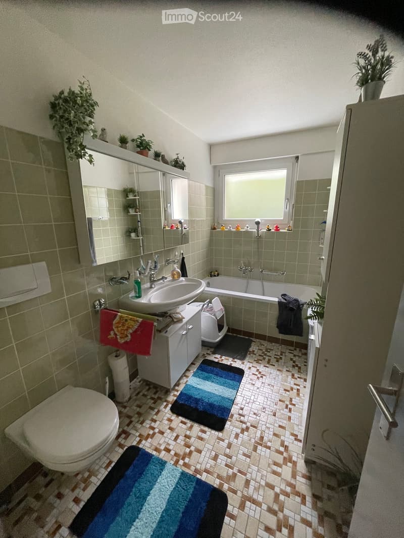 Bathroom with a toilet, sink, bathtub, mirror, and cabinet. Features tiled walls and floor, a window, and decorative plants on the shelves.
