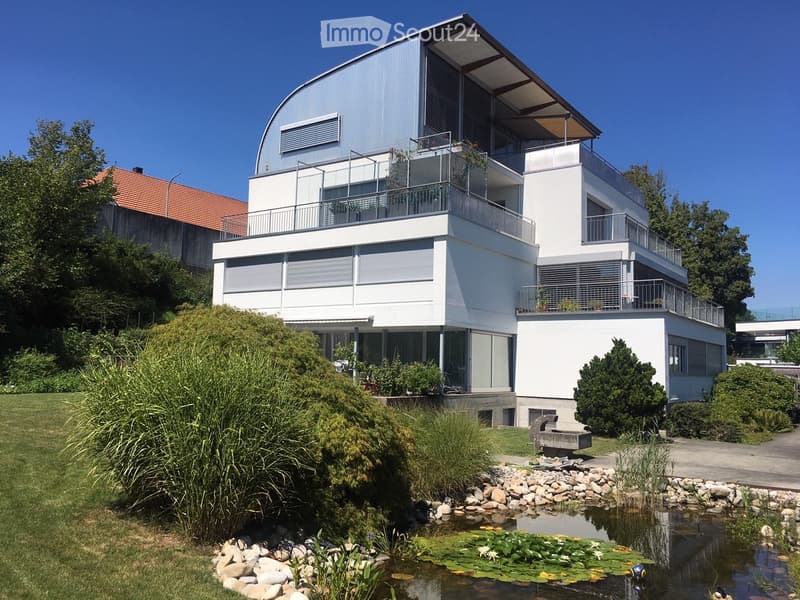 Modern two-story house with white exterior, curved roof, large windows, and balconies. The surrounding area includes a pond with lily pads, a stone bench, and landscaped greenery.