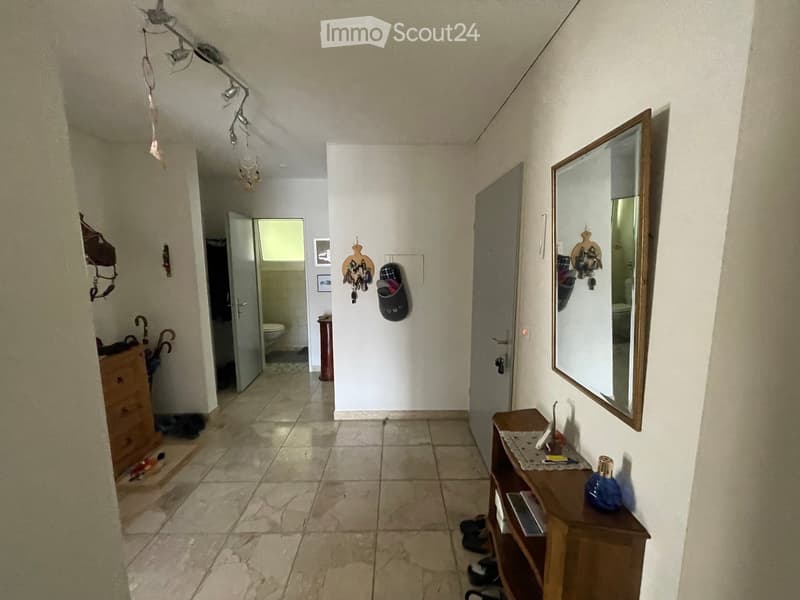 A narrow hallway with tiled flooring, white walls, and several doors leading to different rooms. There is a small wooden table with a mirror, a few items on it, and a coat rack with shoes and bags.