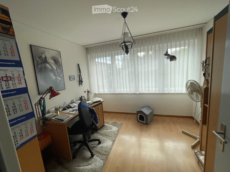 A furnished room with a wooden floor, a desk with a chair, a lamp, a fan, a pet house, a calendar, and a picture frame on the wall.