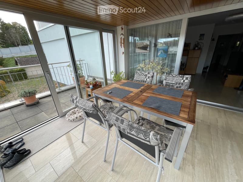 Balcony with a wooden table, two chairs, and a small side table with potted plants. The balcony has large glass doors leading to the interior and a view of the garden outside.