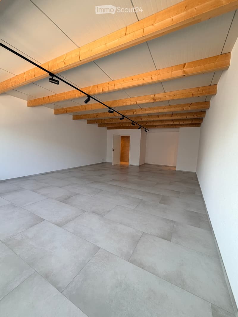 Empty room with exposed wooden beams on the ceiling, grey tiled floor, white walls, and a small doorway leading to another space.