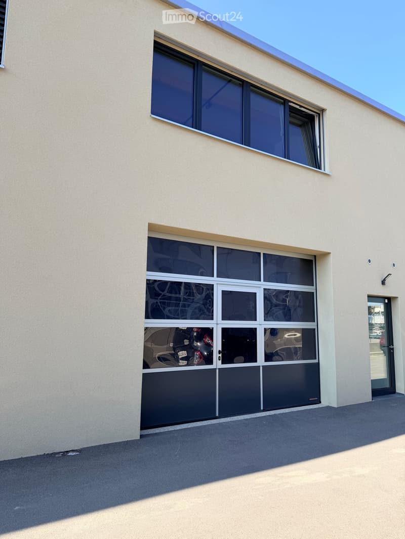 Modern building exterior with large windows and a garage entrance. The facade is light beige with a flat roof and a small overhang above the windows.