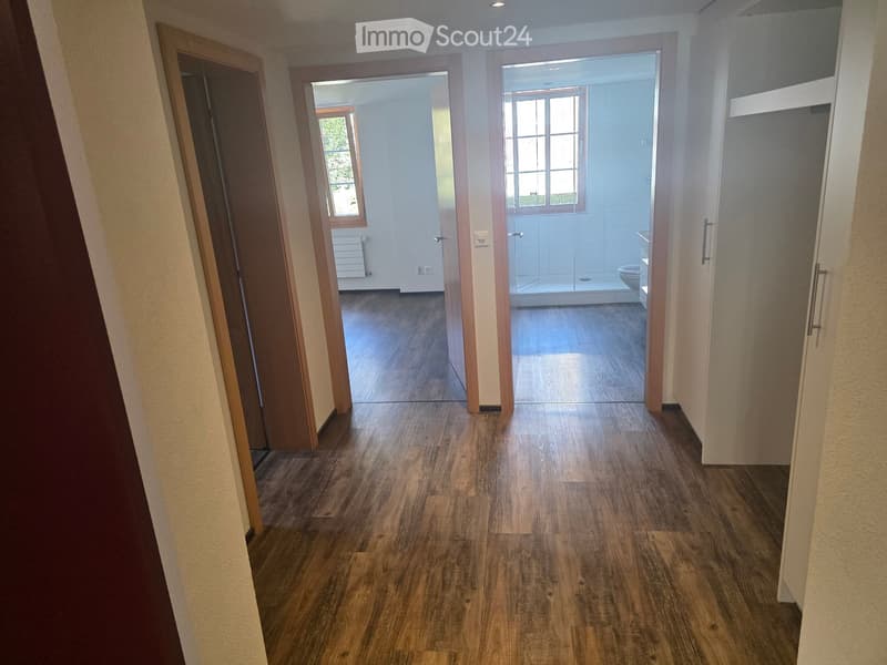 Empty hallway with wooden flooring, leading to a bathroom and another room with large windows and a radiator.