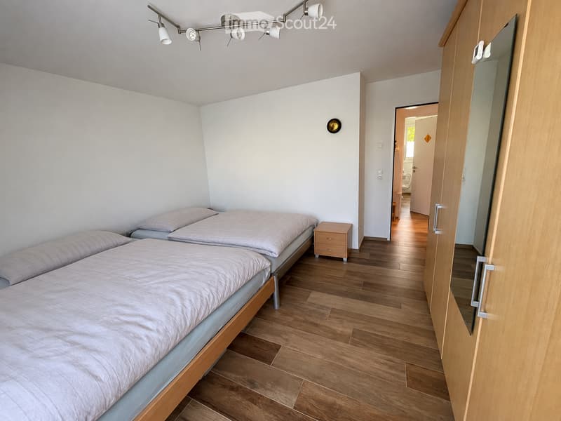 Cozy bedroom with two single beds, wooden nightstand, and a large mirror on the wardrobe. The room features wooden flooring and modern lighting.