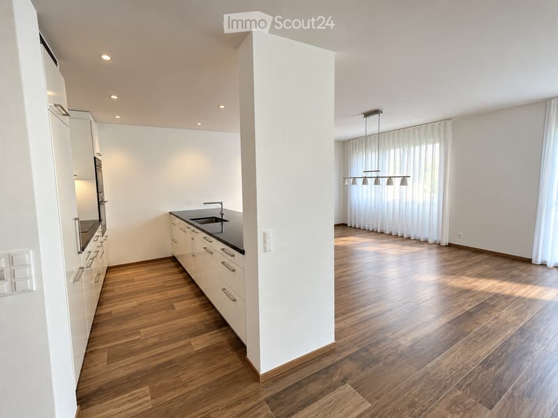 Open-plan kitchen and living area with light wooden flooring, white cabinetry, and large windows with white curtains. The kitchen features a sink, countertop, and built-in appliances, while the living area has a pendant light fixture and ample natural lig