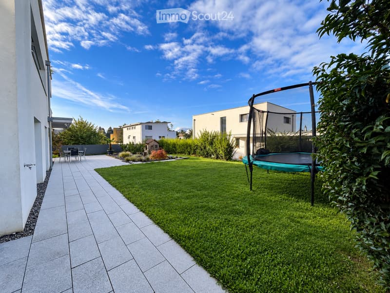 Well-maintained garden with a tiled pathway, green lawn, trampoline, small seating area, and surrounding trees and shrubs.