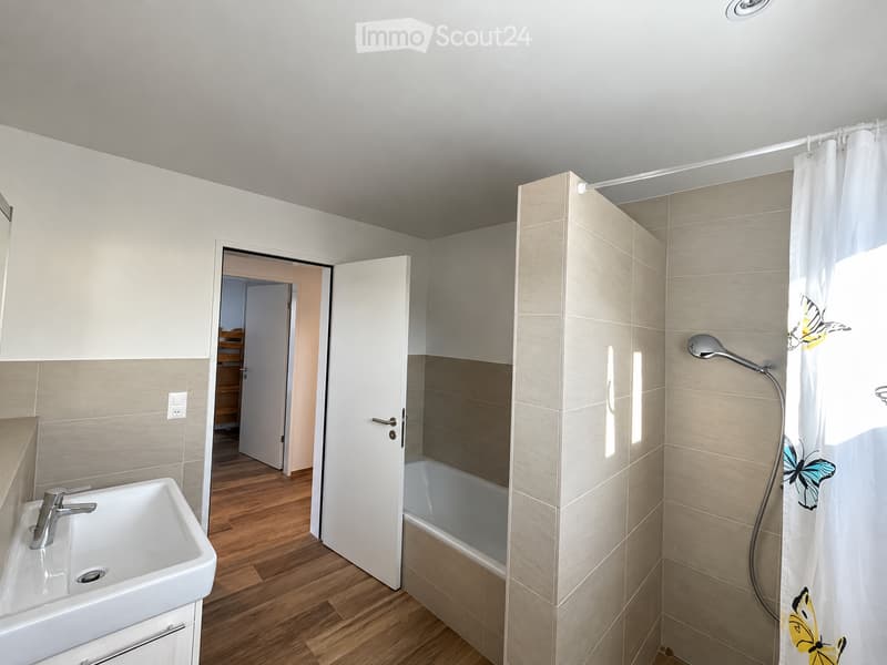 Modern bathroom with a white sink, bathtub, and shower area with a decorative curtain. The room features light-colored tiles and wooden flooring.