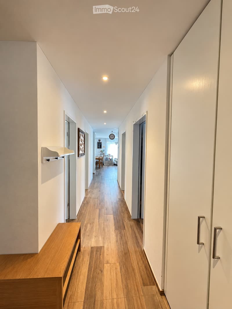 A well-lit hallway with wooden flooring, white walls, and several doors leading to different rooms. A small wooden bench is placed on the left side, and a wall-mounted shelf is visible.