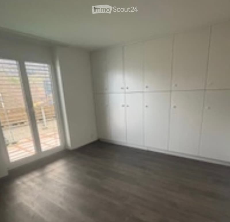 Empty room with white built-in wardrobes, dark flooring, and large windows allowing natural light.