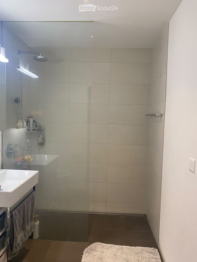 Modern bathroom with a white sink, glass-enclosed shower, beige tiles, and a towel on the floor.