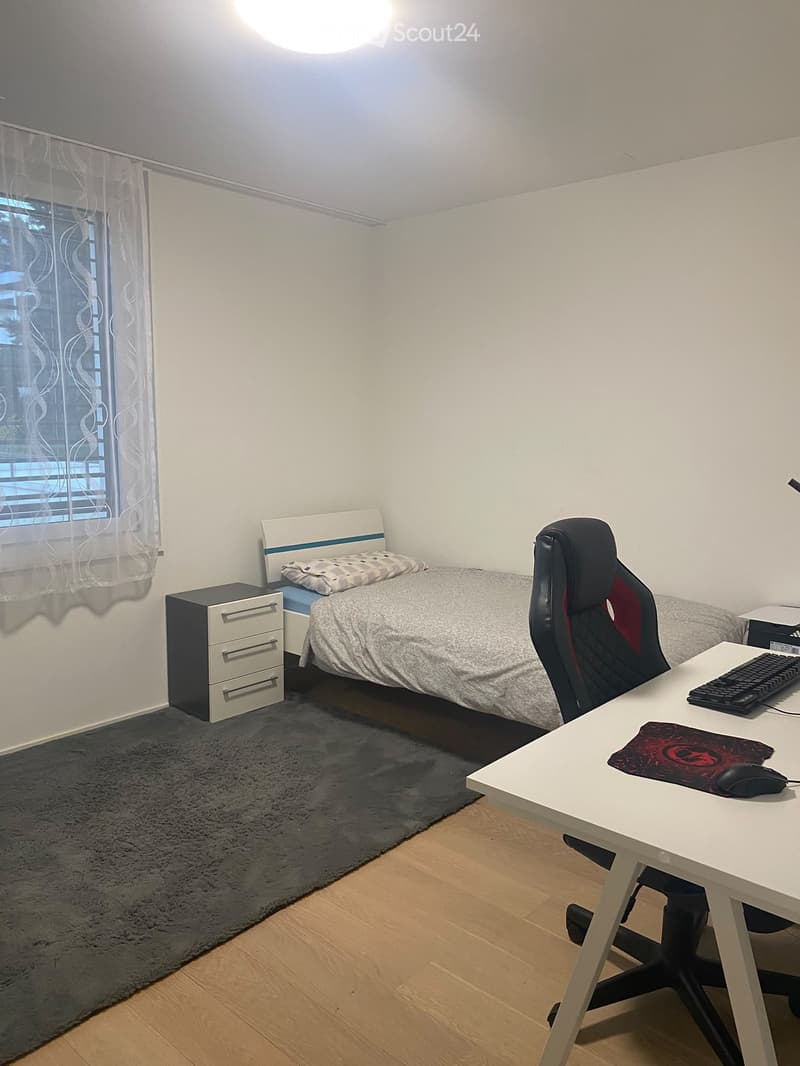 A small, well-lit bedroom with a single bed, desk, and chair. The room features a nightstand, a window with a curtain, and a dark rug on the floor.