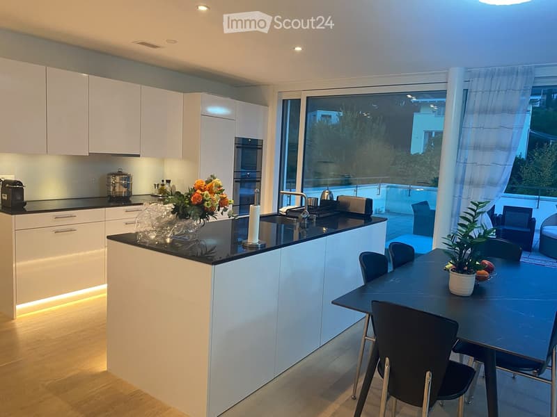 Modern kitchen with white cabinets, black countertops, and a central island. Features a sink, faucet, and a dining area with a table and chairs. A vase with flowers and a bowl of fruit are on the table. The kitchen has built-in lighting and a large window